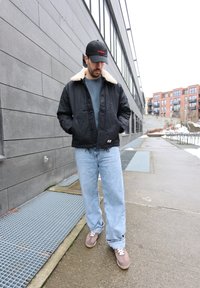 Homme portant une veste noire avec un col blanc, un jean bleu et une casquette noire, debout sur un trottoir en ville près d'un bâtiment gris.