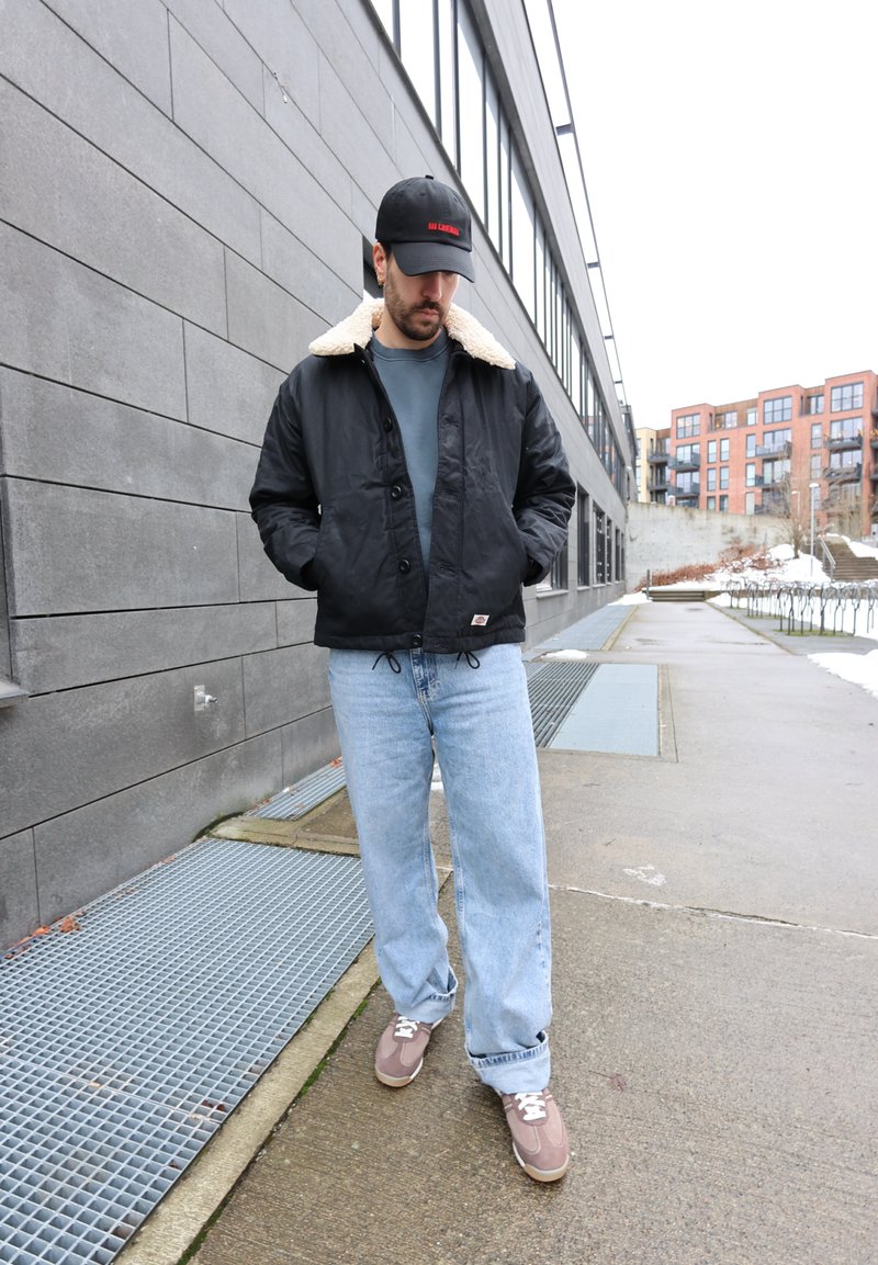 Homme portant une veste noire à col blanc, un jean bleu et une casquette noire, debout sur un trottoir en ville près d'un bâtiment gris.