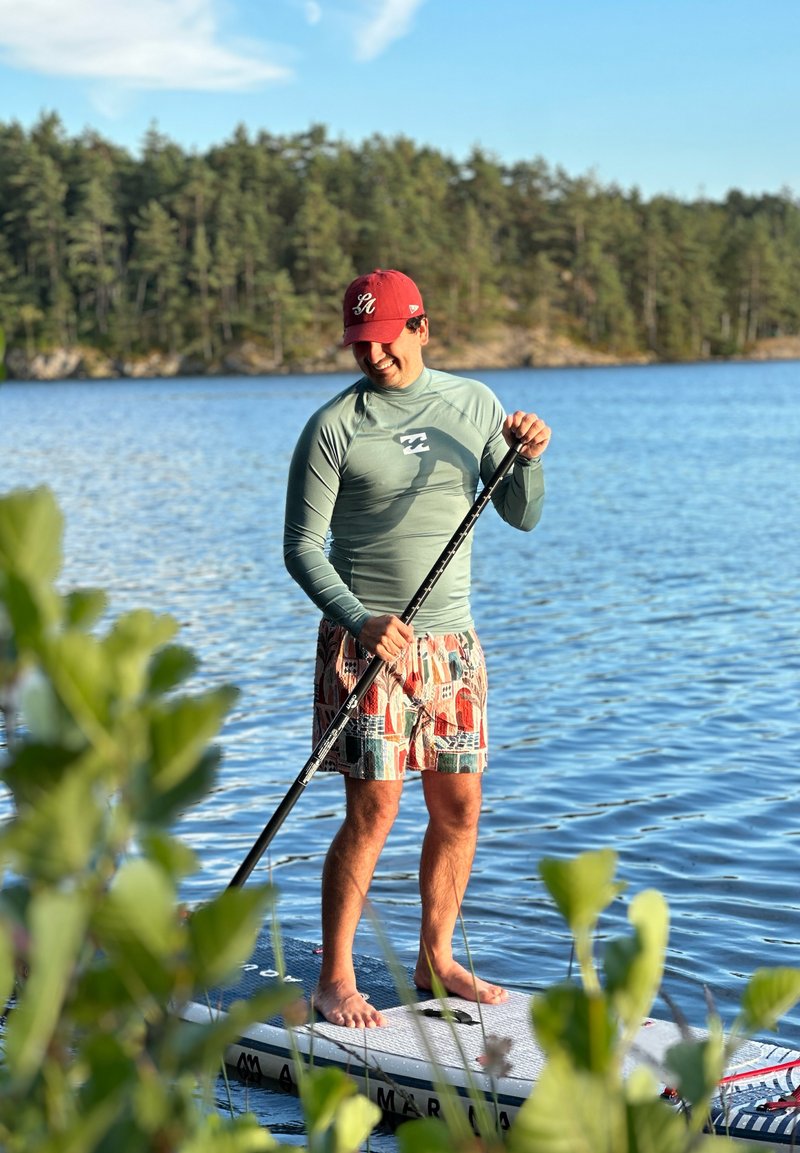 Man wearing a red cap and rash guard paddleboarding barefoot on a calm lake with forested shore in the background.