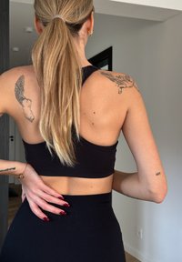 Černý sportovní crop top s designem na zadních popruzích, hladkou texturou a nízkým výstřihem na zádech. Dlouhé rovné vlasy svázané dozadu, na neutrálním pozadí.