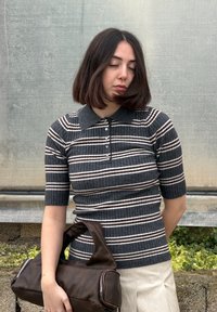 Jeune femme aux yeux fermés portant un polo en tricot rayé gris foncé et une jupe crème, tenant un grand sac à main marron foncé à côté d'elle.