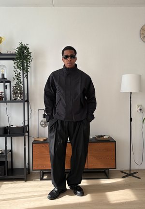 Hombre con chaqueta negra, pantalones negros holgados y gafas de sol, de pie en una habitación minimalista con estanterías, un armario y una lámpara de pie.