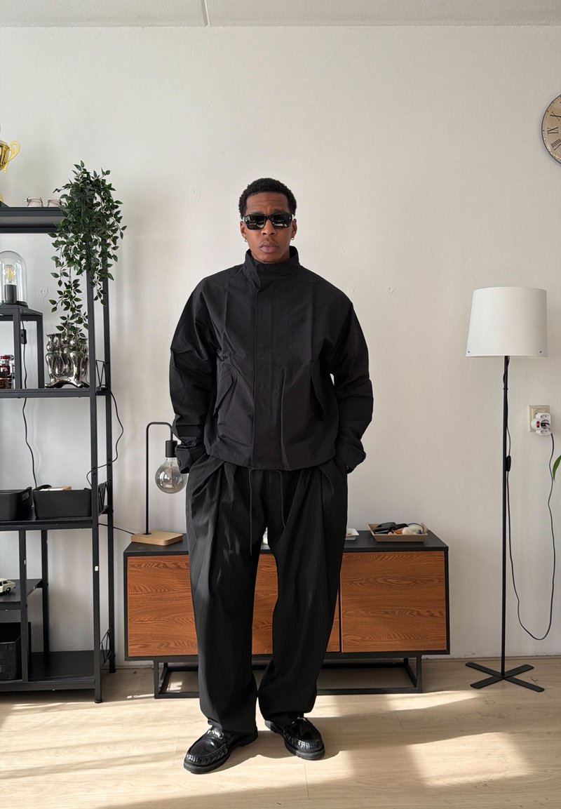 Homme portant une veste noire, un pantalon noir ample et des lunettes de soleil, debout dans une pièce minimaliste avec des étagères, une armoire et une lampe sur pied.