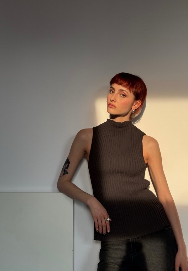 Donna con capelli corti e rossi che indossa una canottiera nera a coste si appoggia a un muro, la luce del sole proietta ombre su di lei e sullo sfondo.