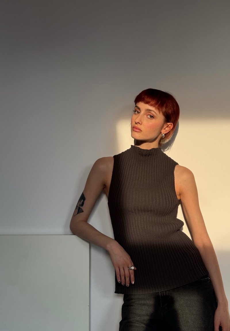 Femme aux cheveux courts et roux portant un haut noir côtelé sans manches s'appuie contre un mur, le soleil projette des ombres sur elle et le fond.