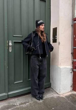 Veste noire courte portée sur un pull noir, associée à un jean noir ample et des chaussures noires à bout pointu. Porte grise en arrière-plan.