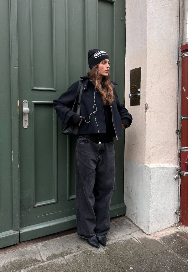 Black cropped jacket over a black sweater, teamed with wide-leg grey jeans and pointed black shoes. Wearing a black beanie with white accents.