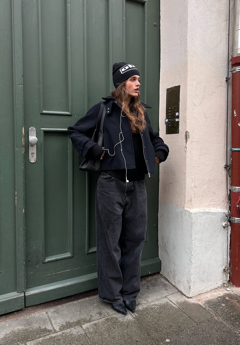 Veste noire courte sur un pull noir, associée à un jean gris à jambes larges et des chaussures noires à bouts pointus. Porte un bonnet noir avec des accents blancs.