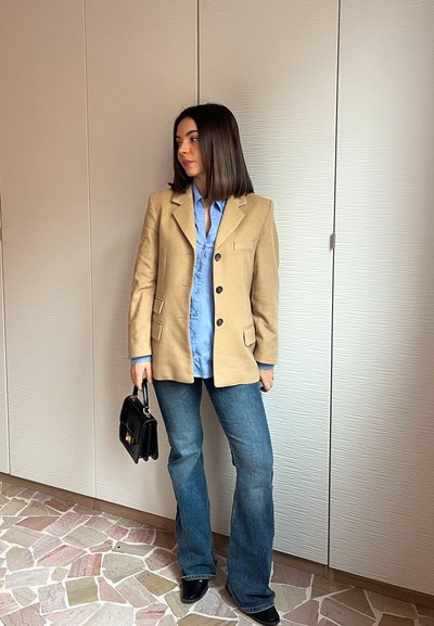 Mujer de pie en el interior, vestida con un blazer beige, camisa azul, vaqueros azules, zapatos negros y sosteniendo un bolso negro, mirando hacia un lado.