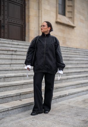 Persona con chaqueta y pantalones negros de gran tamaño, llevando una bolsa negra, de pie en escalones de piedra cerca de un edificio grande con una puerta de madera.
