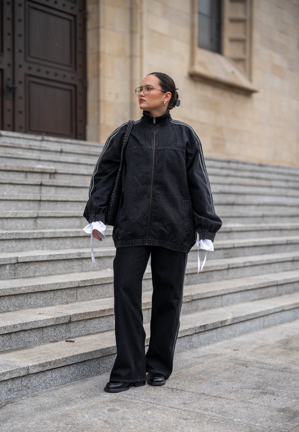 Osoba v čiernej nadrozmernej bunde a nohaviciach, nesúca čiernu tašku, stojaca na kamenných schodoch pri veľkej budove s drevenými dverami.