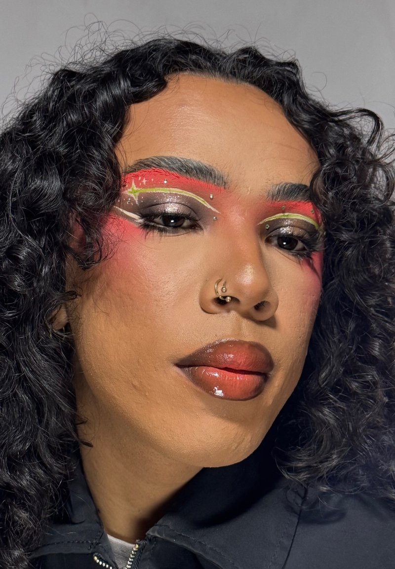 Persona con capelli ricci scuri che indossa un trucco occhi audace rosso e metallico con accenti bianchi e gialli e labbra rosse lucide, con indosso una giacca scura.
