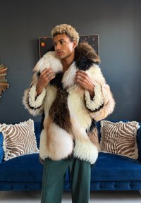 Personne ajustant un grand manteau en fourrure multicolore, debout devant un canapé bleu avec deux coussins à motif zèbre contre un mur sombre.