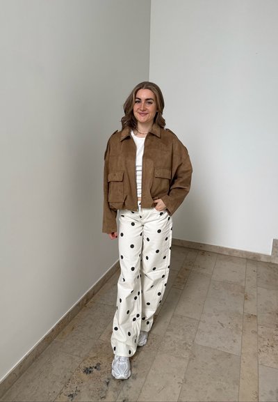 Joven mujer de pie en el interior, con chaqueta marrón, top blanco, pantalones blancos con grandes lunares negros y zapatillas blancas.