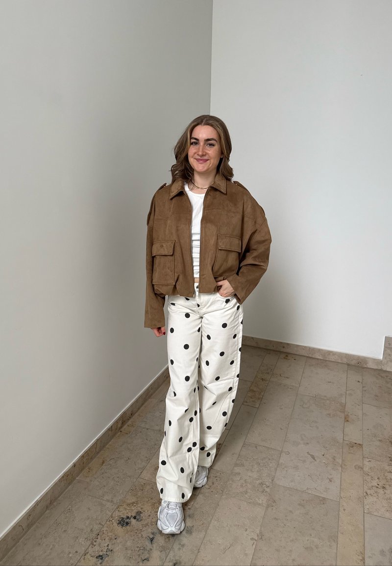 Junge Frau steht drinnen, trägt eine braune Jacke, ein weisses Top, weisse Hosen mit grossen schwarzen Punkten und weisse Sneakers.