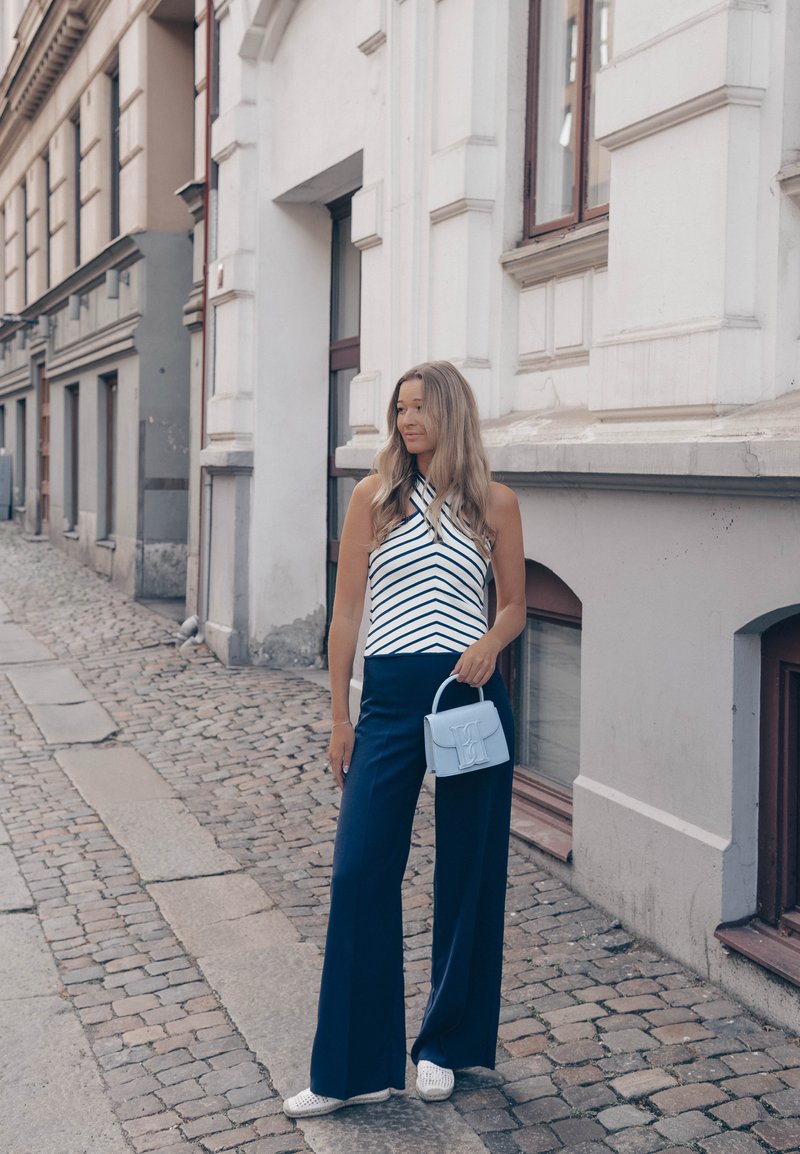 Mujer con pelo largo de pie en calle empedrada, vestida con camiseta de rayas, pantalones anchos oscuros, zapatos blancos y sosteniendo un bolso pequeño azul claro.