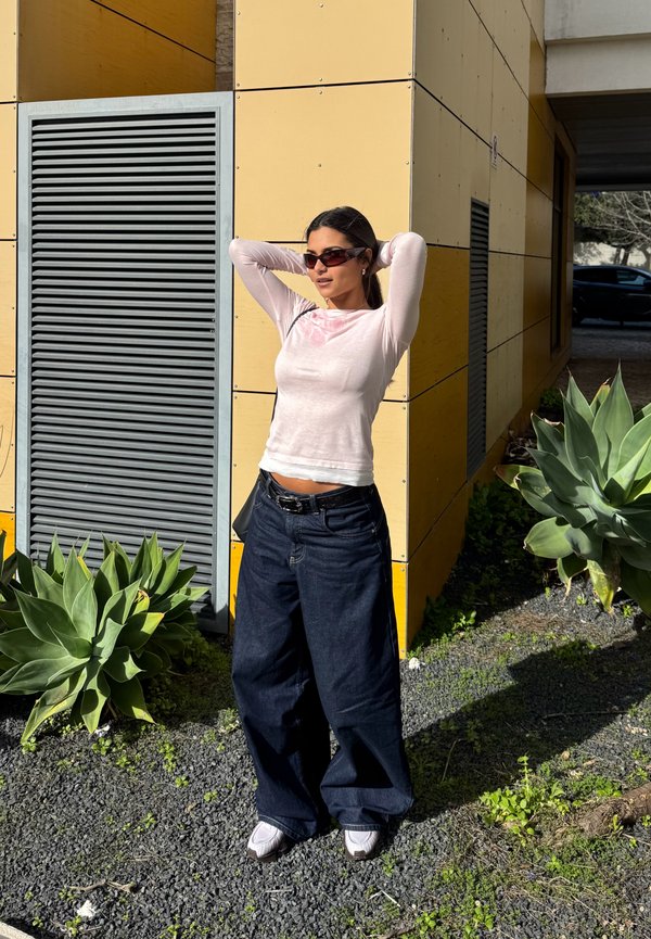 Kvinde med solbriller iført en lyserød langærmet top og løstsiddende mørke jeans står med hænderne bag hovedet nær en gul bygning og grønne planter.