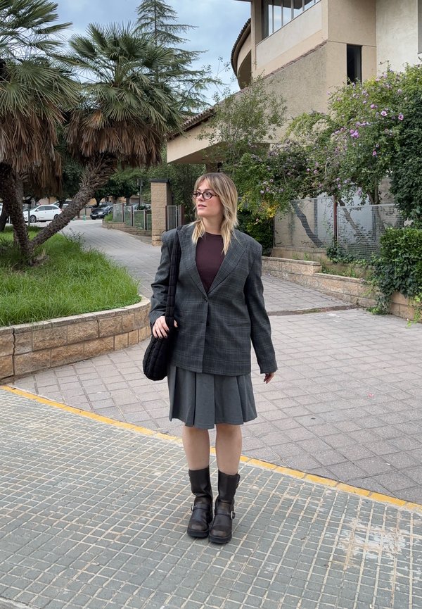 Un sacou oversized cu carouri gri peste un top închis, asortat cu o fustă plisată gri și cizme negre până la gleznă. Se poartă o geantă de umăr neagră.