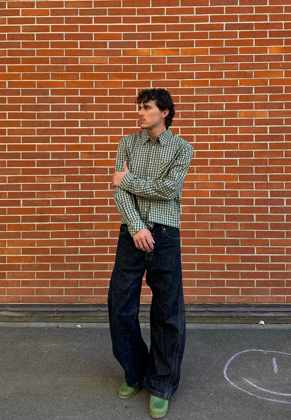 Jeune homme portant une chemise à carreaux verte, un pantalon noir large et des chaussures vertes, debout contre un mur en briques rouges avec un smiley au sol dessiné à la craie à proximité.