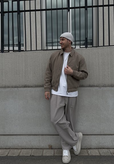 Hombre con chaqueta beige, camisa blanca, gorro gris, pantalones grises holgados y zapatillas apoyado contra una pared de concreto con barras metálicas negras encima.