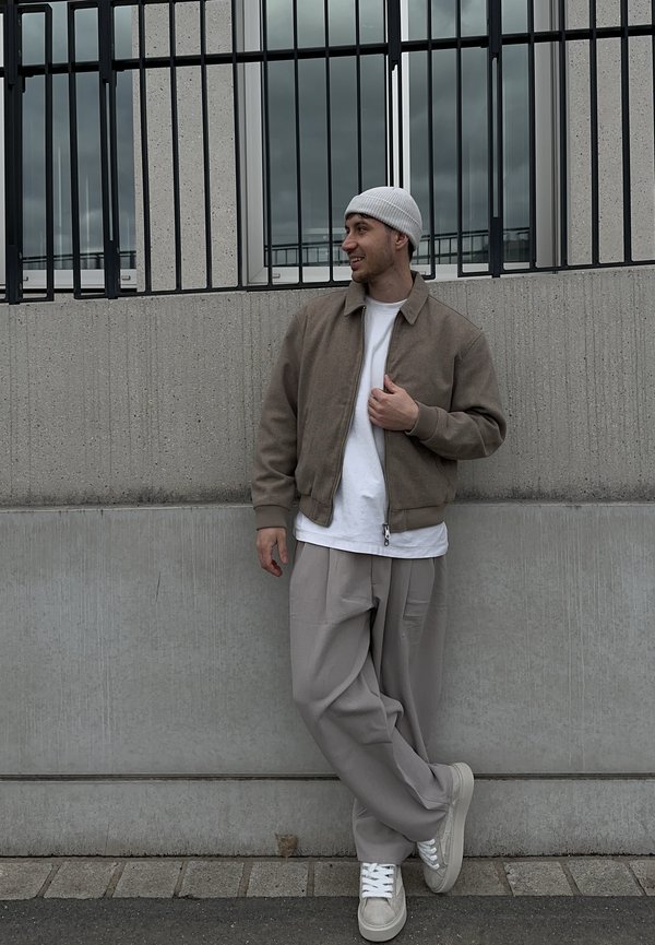 Uomo con giacca beige, camicia bianca, berretto grigio, pantaloni grigi larghi e scarpe da ginnastica appoggiato a un muro di cemento con sopra delle barre metalliche nere.