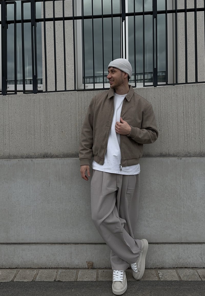 Bărbat în jachetă bej, cămașă albă, căciulă gri, pantaloni largi gri și adidași, sprijinit de un perete din beton cu bare metalice negre deasupra.