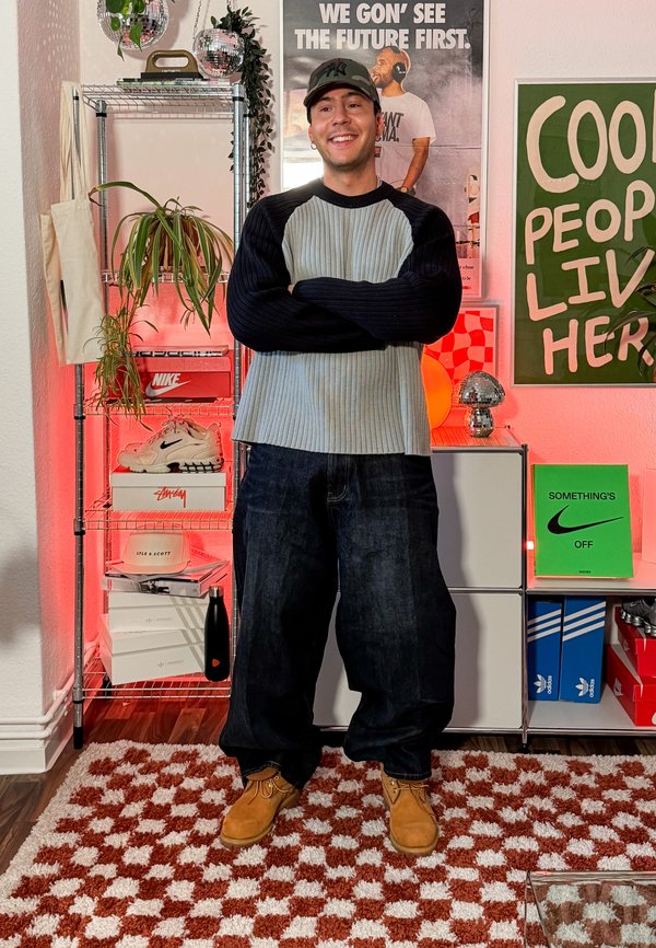 Man i grå och svart tröja, kamouflagemössa och beige skor står och ler i ett rum med sneakers, växter och affischer.