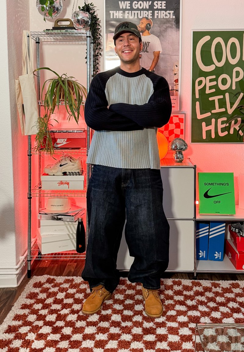 Homme portant un pull gris et noir, une casquette camouflage et des chaussures beige debout en souriant dans une pièce avec des baskets, des plantes et des affiches.