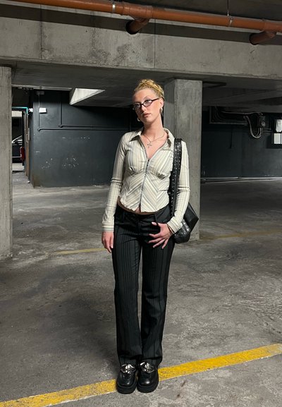 Camisa de botones a rayas con corte slim, pantalones negros a rayas, zapatos negros gruesos y un bolso con tachuelas en un garaje de concreto.