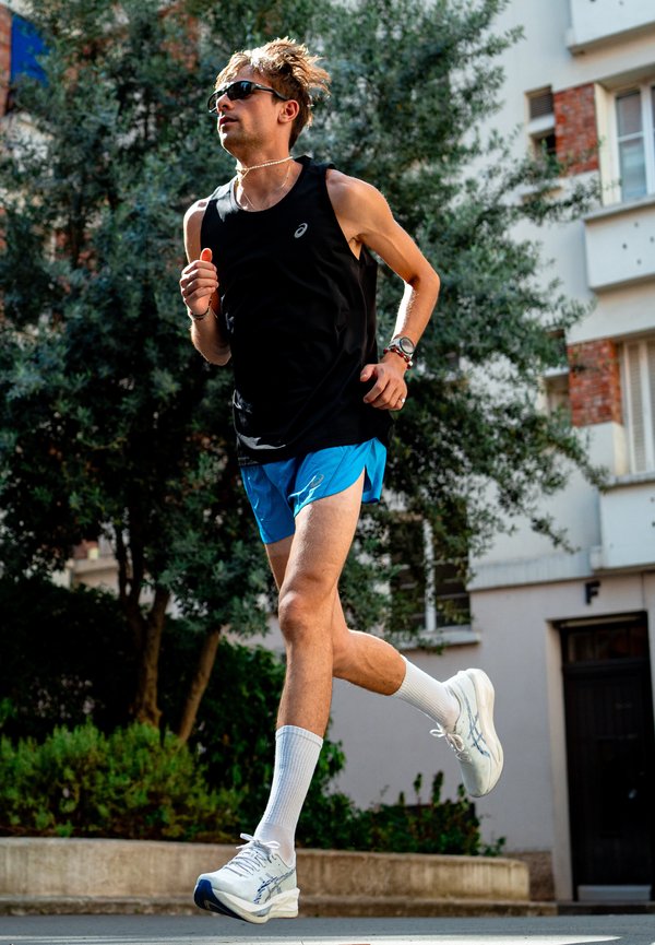 Ung mand løber udendørs iført solbriller, sort tanktop, blå shorts, hvide sokker og løbesko i et grønt urbant område.