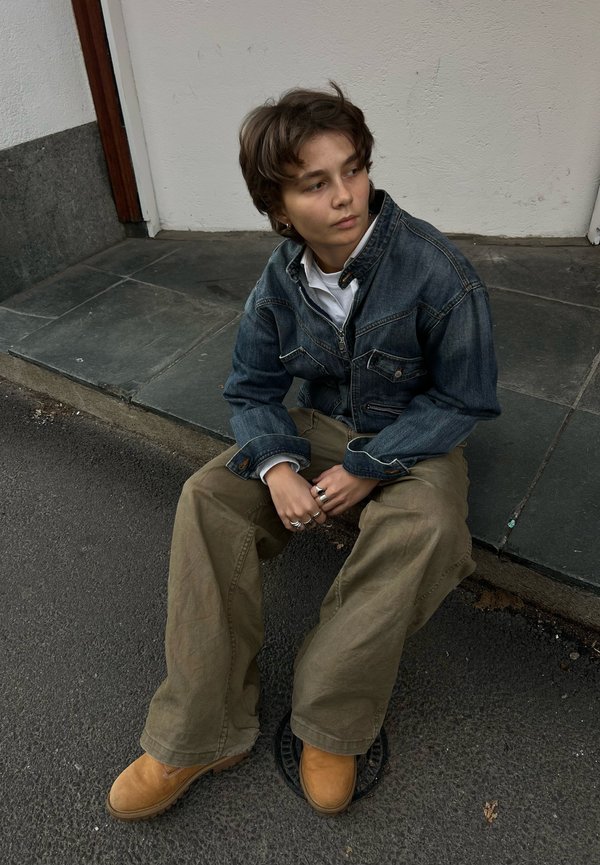 Persona con cabello corto que lleva una chaqueta vaquera, pantalones anchos color caqui y botas marrones sentada en un escalón de piedra mirando hacia un lado.