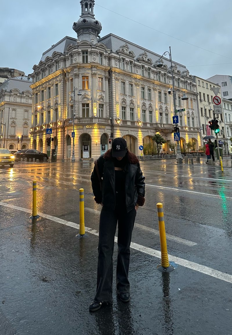 Μαύρο δερμάτινο μπουφάν με γούνινες λεπτομέρειες, μαύρη μπλούζα και σκούρο παντελόνι· στέκεται σε βρεγμένο δρόμο κοντά σε ιστορικό κτίριο με ζεστό φωτισμό.