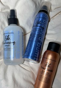 Trois produits capillaires disposés sur un tissu clair : un flacon vaporisateur transparent, une bombe de mousse bleue brillante et un flacon de protection thermique cuivré bronzé.