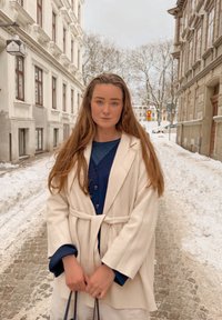 Νεαρή γυναίκα με μπεζ παλτό στέκεται σε ένα χιονισμένο λιθόστρωτο δρόμο μεταξύ κτιρίων ευρωπαϊκού τύπου το χειμώνα.