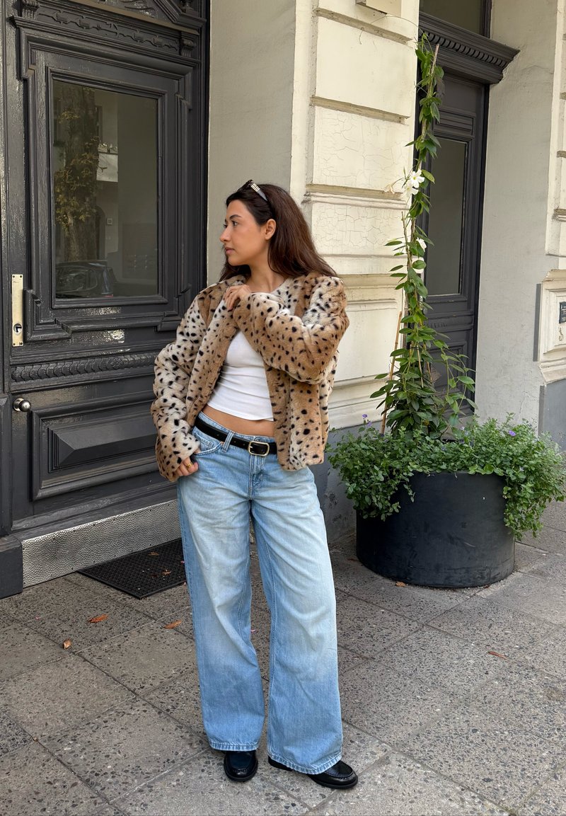Jeune femme portant une veste en fourrure tachetée, un crop top blanc, un jean large bleu et des chaussures noires, se tenant près d'une porte sombre et d'un grand pot de plante sur le trottoir.