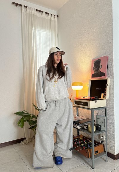 Sudadera gris de gran tamaño y pantalones de chándal de pierna ancha, combinados con una gorra de béisbol. Calzado azul. El fondo presenta una planta, una lámpara y un tocadiscos.