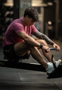 Veslovací stroj s osobou, která ho používá, oblečenou v růžovém tričku a černých šortkách. Pozadí tvoří fitness vybavení a málo osvětlené.