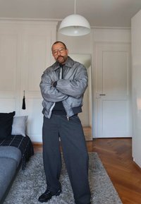 Mees prillide, habeme ja kõrvarõngastega seisab määrdunud kätega toas, kandes halli jopet, tumedaid pükse ja musti vabaajajalatsid hallil vaibal.