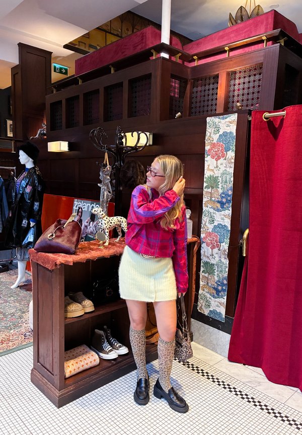 Joven mujer con camisa de cuadros roja, falda crema y botas con estampado de leopardo, de pie dentro de una tienda de estilo vintage con estanterías de madera y cortina roja.