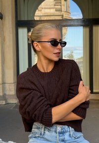 Suéter marrón de punto con puños acanalados, combinado con pantalones cortos de mezclilla de tiro alto en azul claro. Gafas de sol ovaladas negras completan el look.