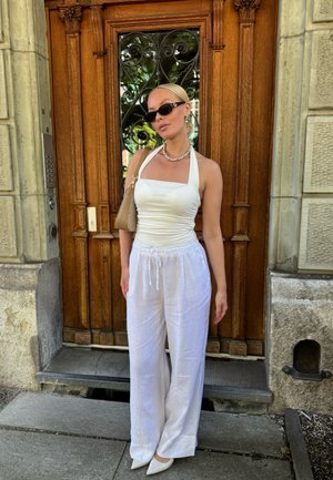 Top blanco de tirantes acompañado de pantalones de lino blancos holgados, adornado con un bolso de color claro y gafas de sol oscuras, frente a una puerta de madera.