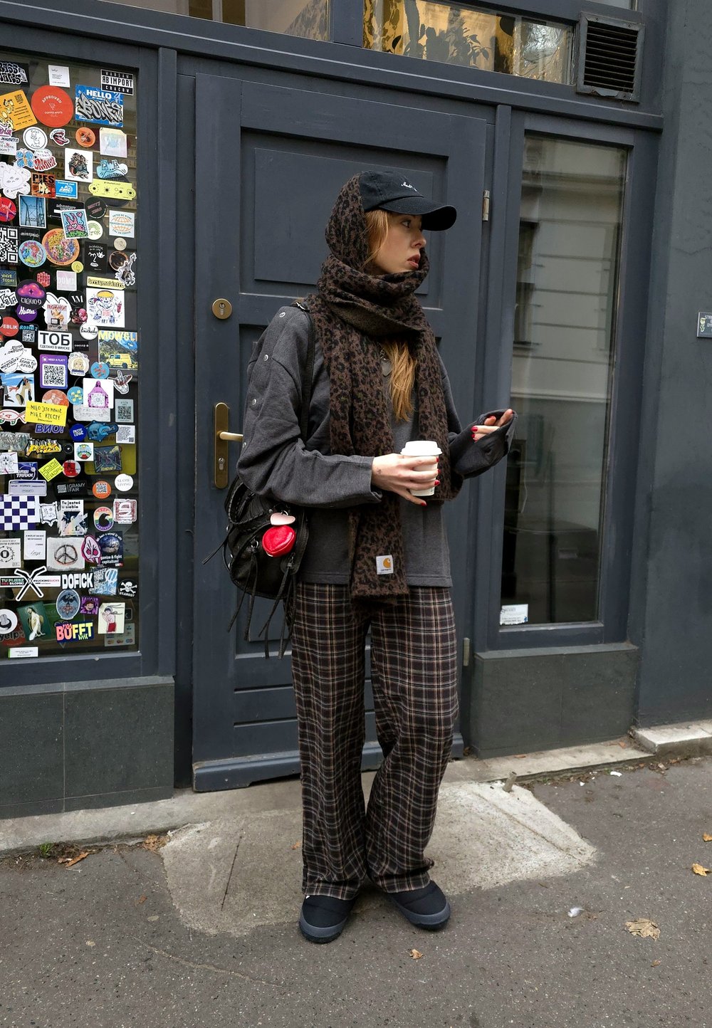 Tumši pelēkas krāsas krekls ar garām piedurknēm, leoparda raksta šalle, rūtainas bikses, melna cepure, turot kafijas krūzi. Melnā soma ar sarkanu aksesuāru. Uz durvīm ir uzlīmes.