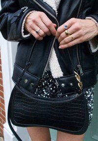 Borsa nera lucida con motivo coccodrillo, dalla forma curvata, con accentuazione in catena d'oro e tracolla regolabile. Indossata con una giacca di pelle e una gonna con paillettes.