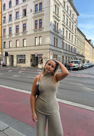 Mono beige brillante con cuello alto y piernas anchas. Textura suave, combinado con un pequeño bolso. Escena urbana de fondo.