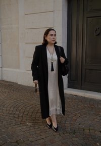 Cappotto lungo nero su un vestito di satin crema con bordo in pizzo. Gli accessori includono una lunga collana con un pon-pon e una piccola borsa nera. Scarpe con tacco nere.