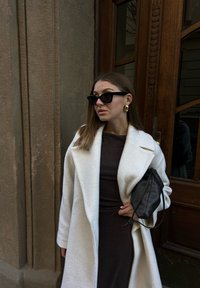 Femme portant un manteau blanc et une robe foncée, tenant un sac à main noir, debout près d'une porte en bois, portant des lunettes de soleil noires et des boucles d'oreilles dorées.