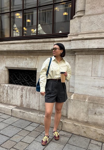 Camisa amarilla a rayas, pantalones cortos grises con pliegues, zapatillas marrones y burdeos, bolso azul, sosteniendo una taza de café, de pie junto a una pared de piedra.