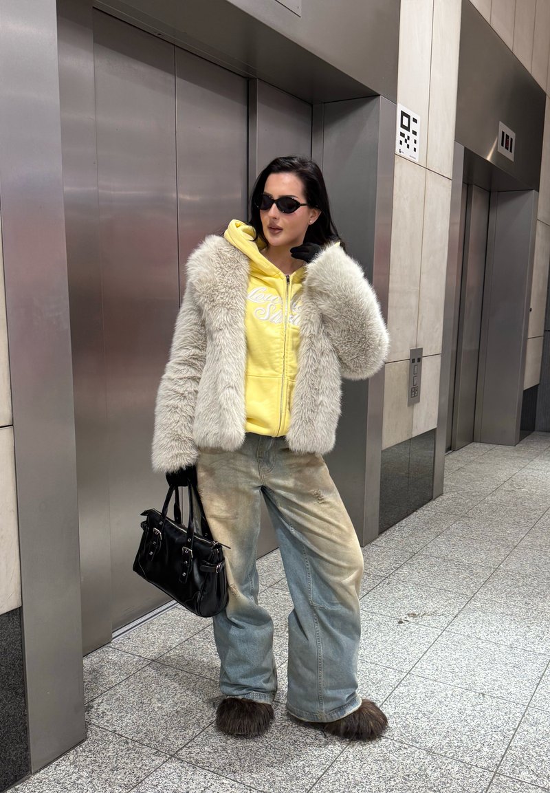 Femme portant des lunettes de soleil, un sweat à capuche jaune, une veste en fourrure grise, un jean large et des chaussures en fourrure, tenant un sac à main noir, debout près de deux ascenseurs fermés.