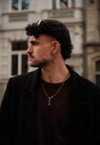 Une personne portant un manteau noir et un pull marron, avec des bijoux dorés incluant un collier chaîne et des boucles d'oreilles. En arrière-plan, un bâtiment.