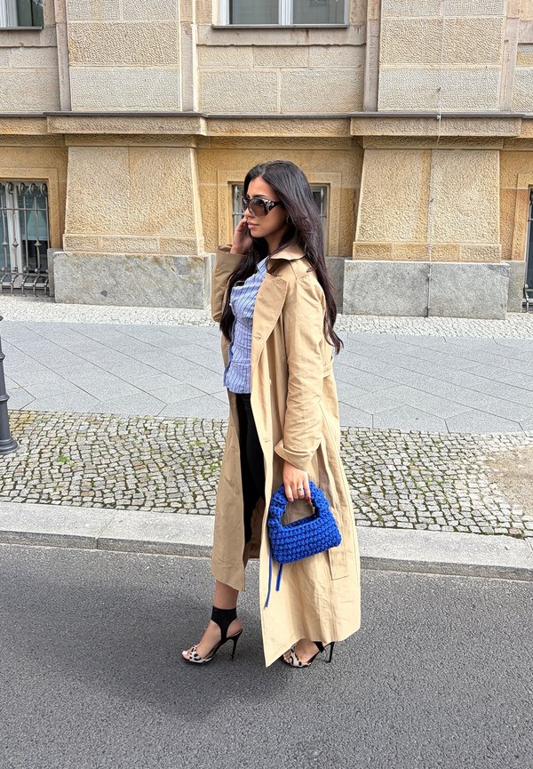 Femme aux longs cheveux foncés portant des lunettes de soleil, un trench beige, une jupe noire et des talons à imprimé léopard, tenant un sac à main bleu dans une rue de la ville.